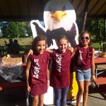 Three children wear Augsburg t-shirts in front of Auggie Eagle standup